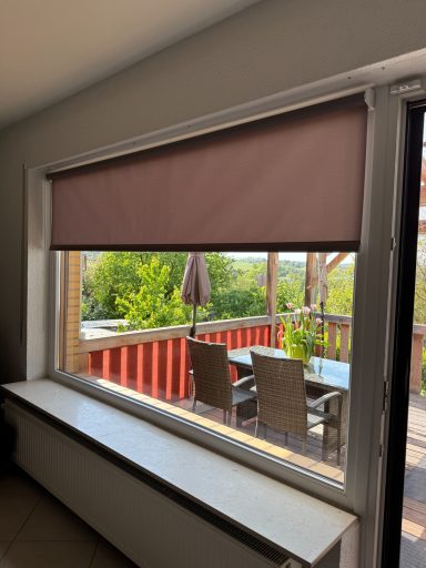Brauner Rolladen vor einem Fenster mit Blick auf eine Terrasse und Gartenmöbel.