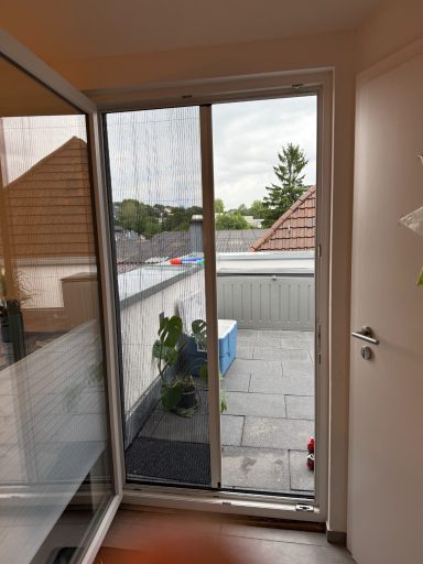 Blick durch eine Glastür auf einen Balkon mit Pflanzen und Dachlandschaft.