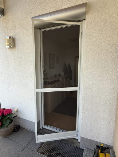 Weiße Türe mit Insektenschutz, öffnend zu einem Innenbereich mit Pflanzen davor.