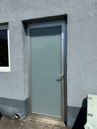 Graue Wand mit einer modernen, grauen Tür und einem Fenster daneben.