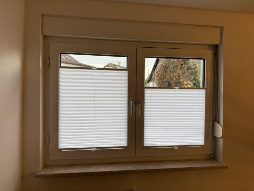 Zwei Fenster mit weißen, gerollten Jalousien und einem hellen Wandbereich.