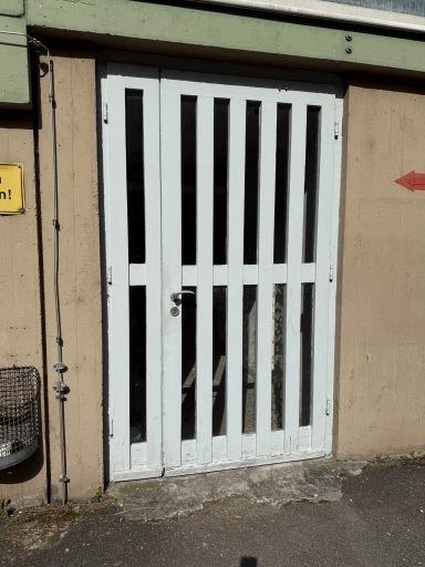 Weiße Gittertür an einer grauen Wand, mit einem Türgriff und Bodenbelag aus Beton.