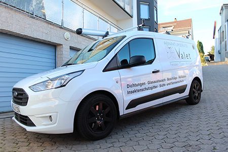Weißer Lieferwagen mit schwarzem Schriftzug, geparkt vor einem Garagentor.
