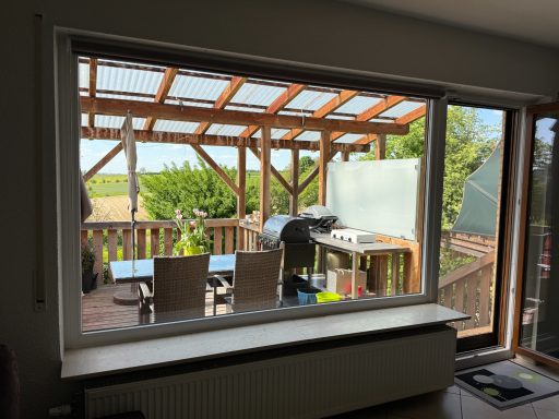 Blick durch ein Fenster auf eine Terrasse mit Grill und Essbereich.